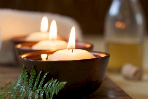 The image shows three lit candles in dark-colored bowls, with a green fern and a bottle in the background, creating a soothing atmosphere.