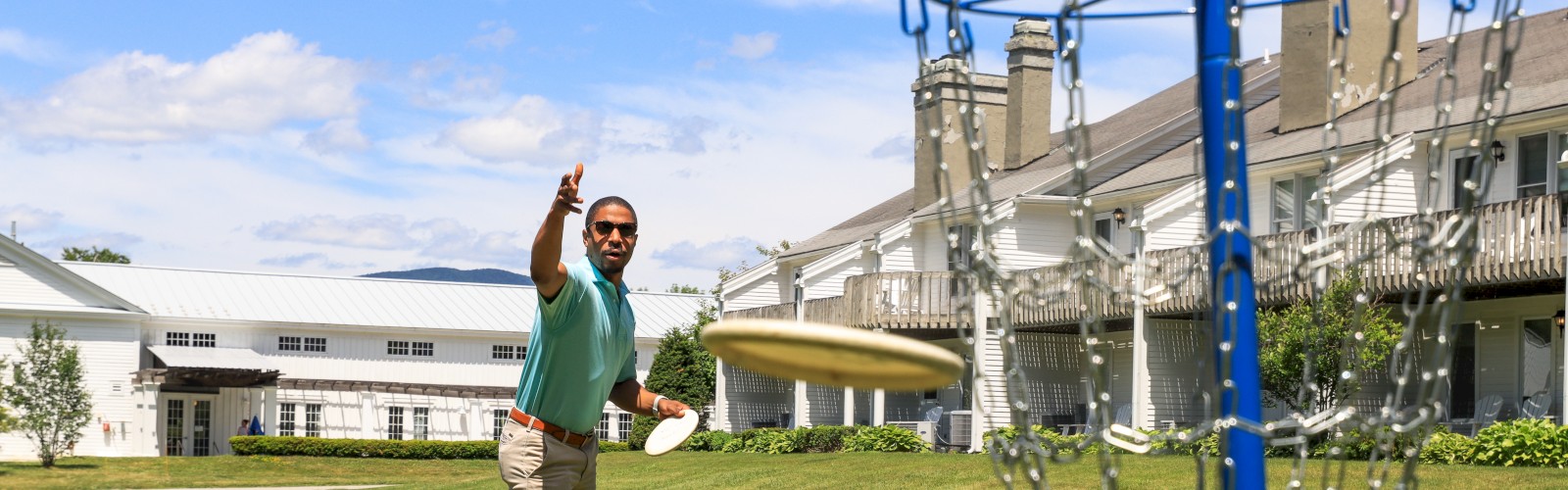 A person is playing disc golf on a grassy lawn with a blue disc golf basket in the foreground and buildings in the background, ending the sentence.