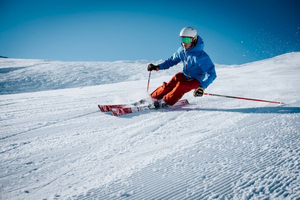A skier in a blue jacket and red pants is making a turn on a groomed snowy slope under a clear blue sky, wearing a helmet and goggles.