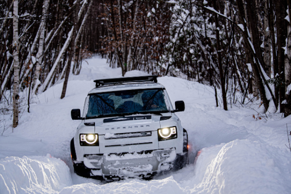 A white SUV is driving through a snow-covered forest path, surrounded by trees with its headlights on.