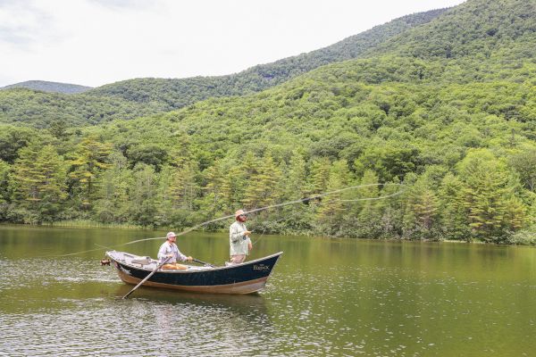 Two people fishing from a small boat on a calm lake surrounded by lush green mountains and forested hills in the background.