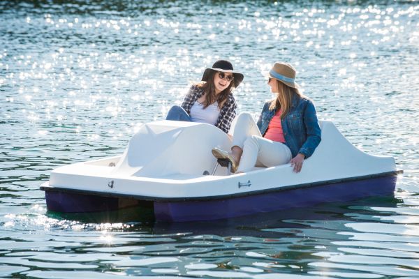 Two women are relaxing and chatting while sitting on a pedal boat in the water, wearing casual clothes and hats, under bright sunlight.