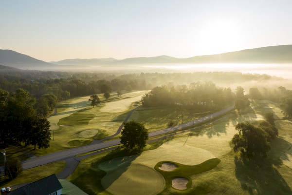 An aerial view of a lush golf course at sunrise, with sand traps, trees, and a winding road, surrounded by mountains and morning fog.
