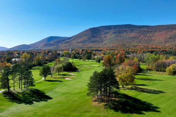 The image features a scenic golf course with lush green fairways, trees, and a backdrop of mountains under a clear blue sky.