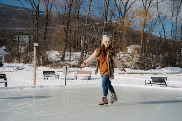 A person is ice skating on an outdoor rink surrounded by snow, trees, and benches on a clear day.