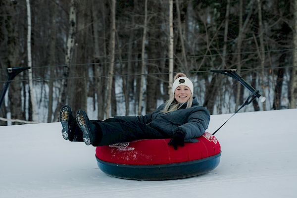 A person is sitting on a red and blue snow tube, smiling, with a snowy forest backdrop. They are dressed warmly, wearing a hat and gloves.