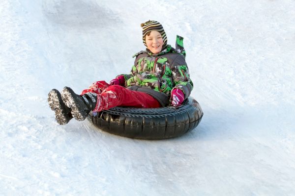 A person wearing winter clothing is sitting on a black inner tube while sliding down a snowy slope, smiling and enjoying the ride.