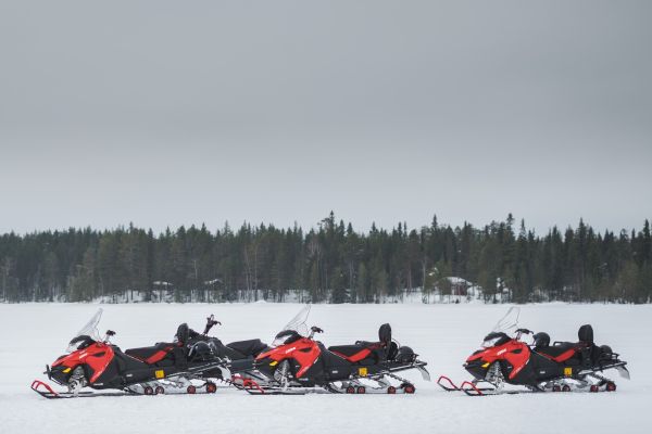 Four red snowmobiles are parked in a snowy landscape with a forest and cloudy sky in the background, suggesting a winter adventure location.
