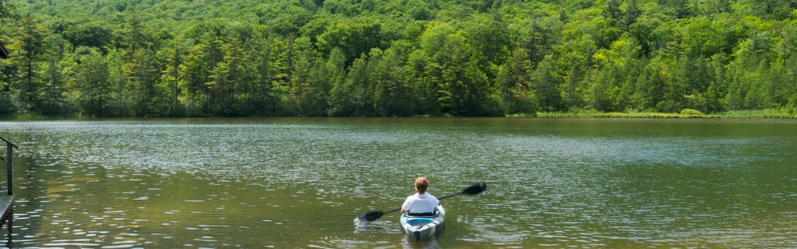 A person is kayaking on a calm lake surrounded by lush green mountains and trees under a partly cloudy sky.