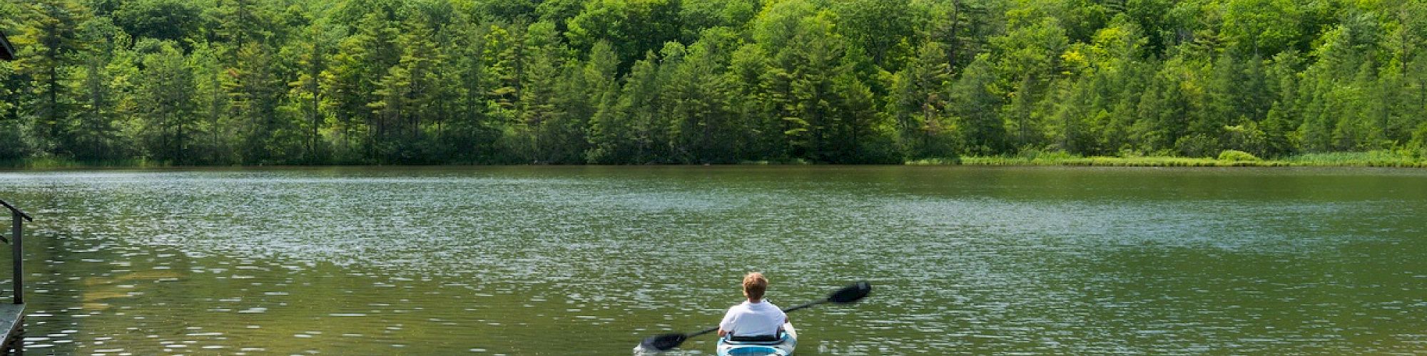 A person is kayaking on a calm lake surrounded by lush green mountains and trees under a partly cloudy sky.