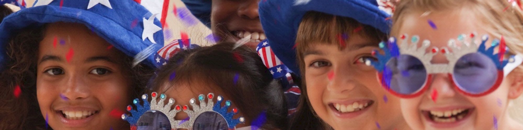 A group of children wearing patriotic hats and sunglasses, decorated with American flag themes, smiling and celebrating.