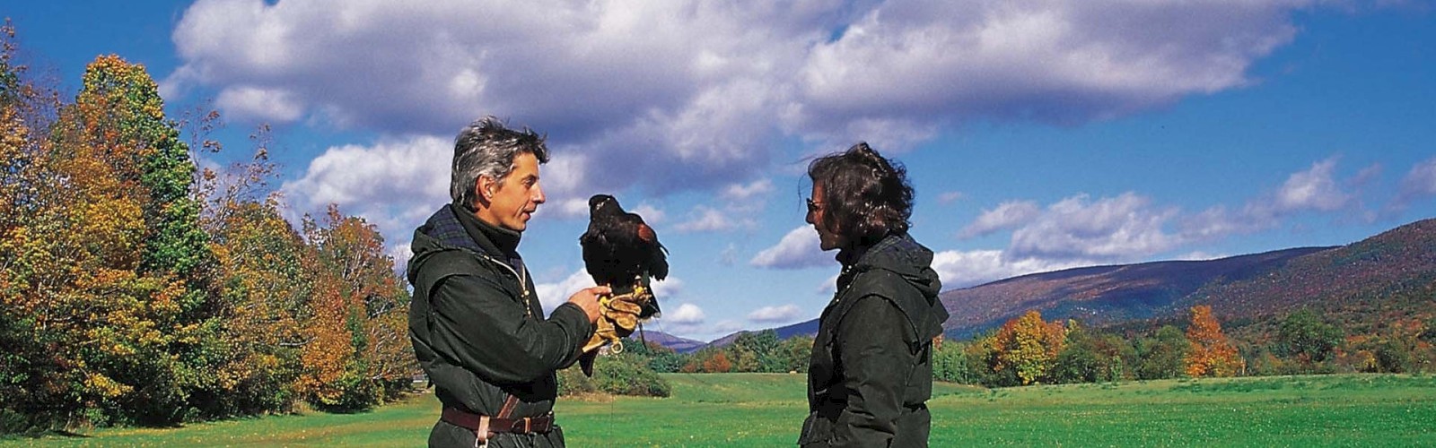 Two people outdoors with a bird on one person's hand, standing on a grassy field with trees and a mountain in the background.