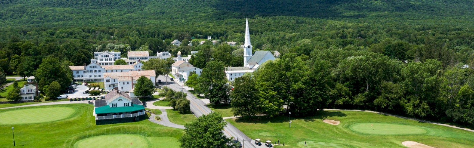 The image features a picturesque village with a white steeple church, surrounded by greenery, a golf course, and mountains in the background, ending the sentence.