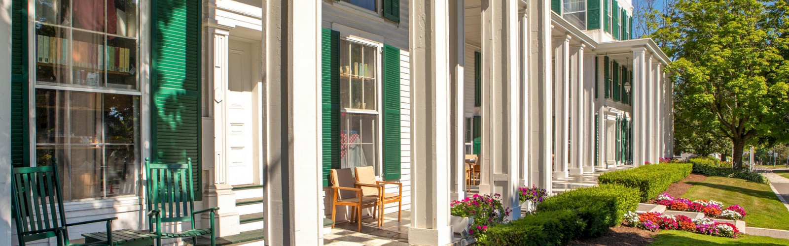 A beautiful building with green shutters, white columns, rocking chairs, and flowers along the walkway, under a clear blue sky.