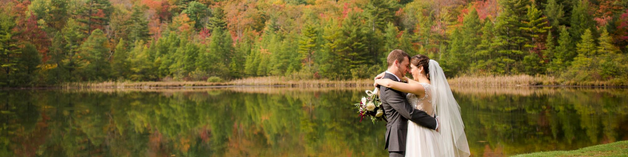 A couple in wedding attire embraces by a serene lake with a backdrop of vibrant, multicolored autumn foliage on a mountainside in the distance.