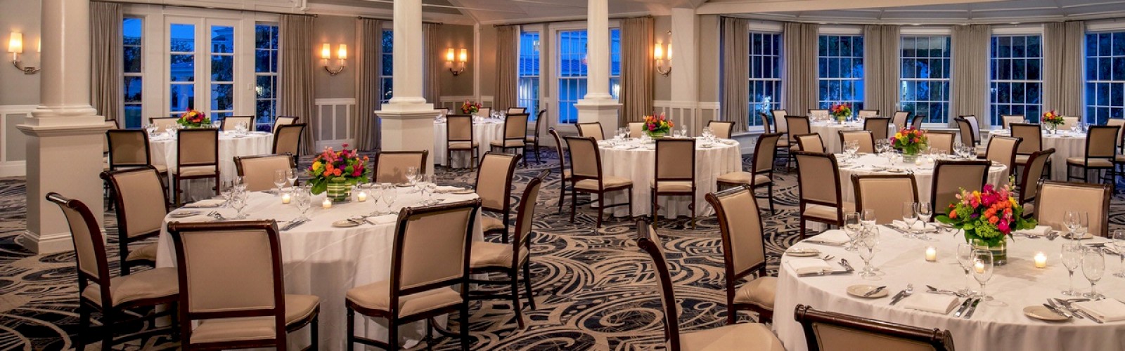 The image shows an elegantly set banquet hall with round tables and chairs, chandeliers, floral centerpieces, and large windows.