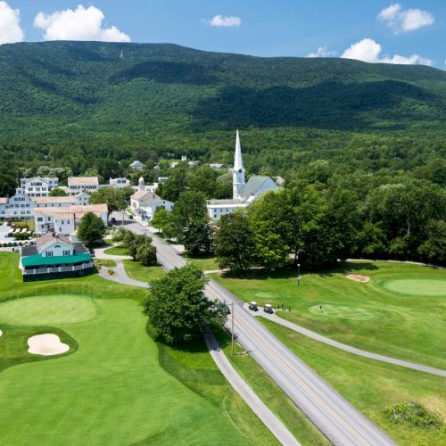 A scenic town nestled in a valley with a white church, surrounded by lush greenery, golf courses, and a mountainous backdrop under a blue sky.