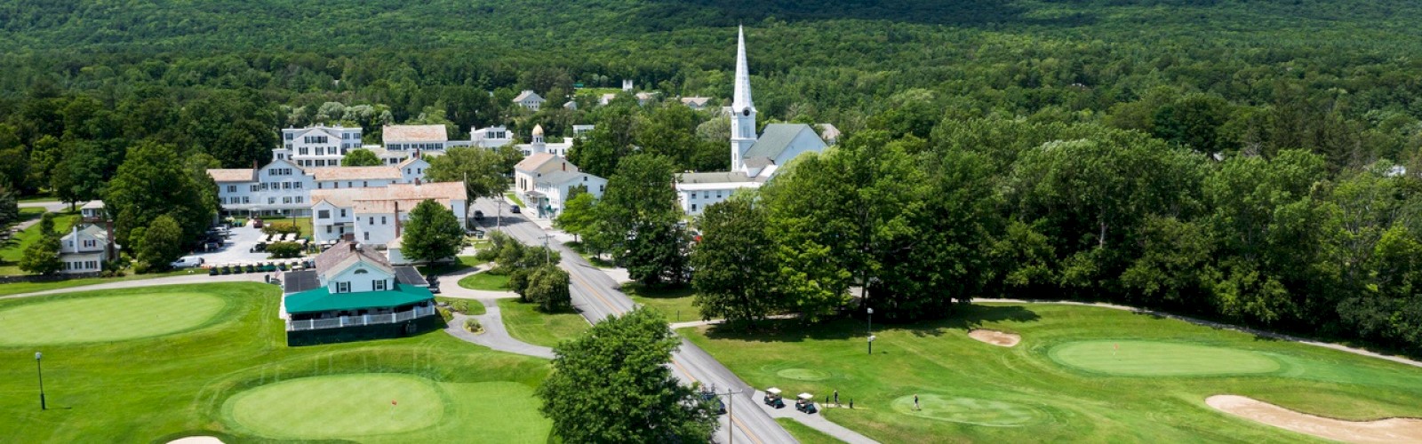 An aerial view of a small town with a church steeple, surrounded by green fields and a golf course, set against a mountain backdrop.
