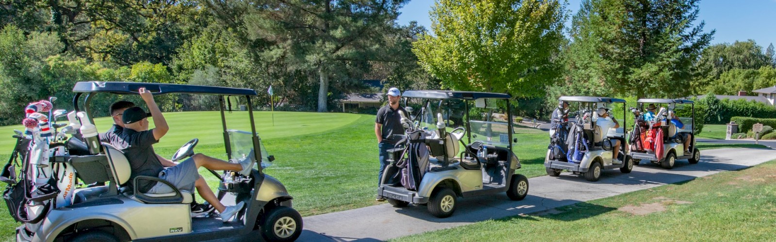 Golfers in golf carts on a path, with golf bags attached to the carts; they are on a golf course surrounded by trees and green grass, ending the sentence.