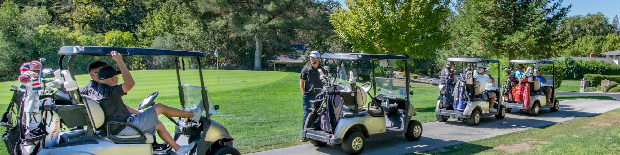 Golfers in golf carts on a path, with golf bags attached to the carts; they are on a golf course surrounded by trees and green grass, ending the sentence.