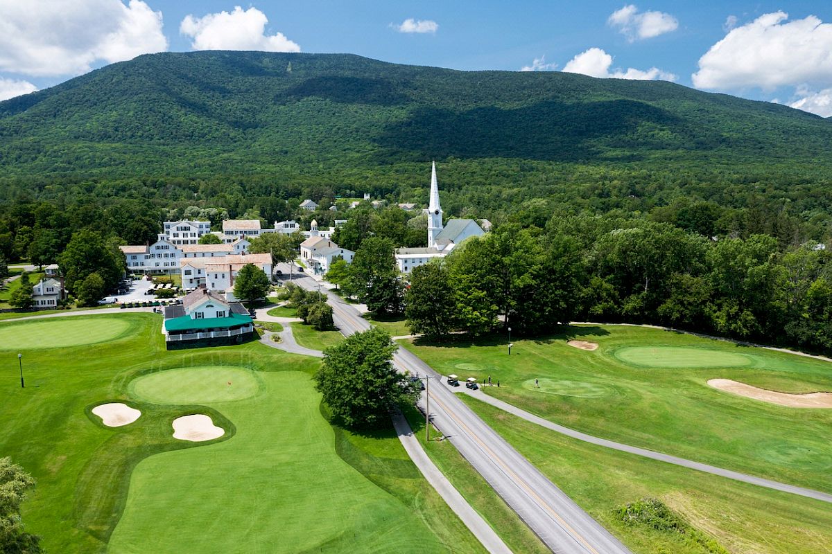 A scenic aerial view of a small town with a church, surrounded by lush greenery, mountains, and a golf course with sand bunkers and roads.