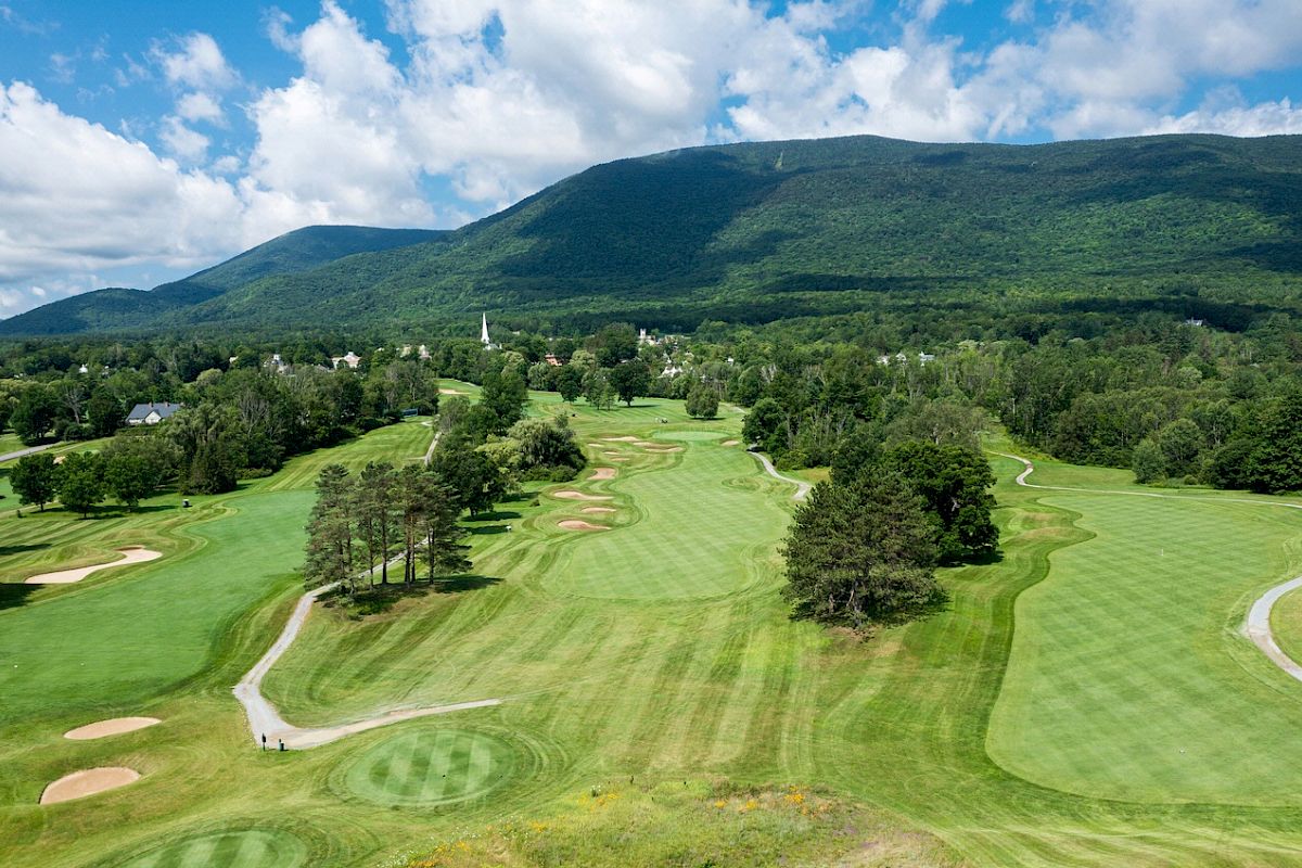 A scenic golf course with well-manicured fairways and greens surrounded by trees, with mountains and a blue sky in the background ending the sentence.