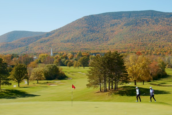 Two people walking on a golf course surrounded by green grass, trees, and hills with autumn foliage under a clear sky.