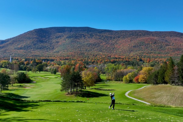 A person is playing golf on a course surrounded by trees with autumn foliage and large hills in the background, under a clear blue sky.