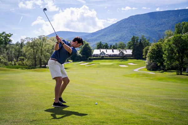 A person is playing golf on a lush green course with mountains and a clubhouse in the background.