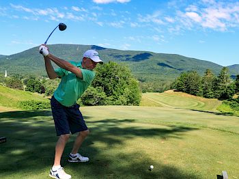 A person prepares to swing a golf club on a scenic golf course with green hills and a blue sky in the background.