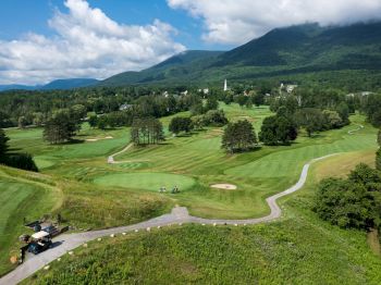 The image shows a scenic golf course with lush green fairways, people playing golf, golf carts, and mountains in the background under a partly cloudy sky.