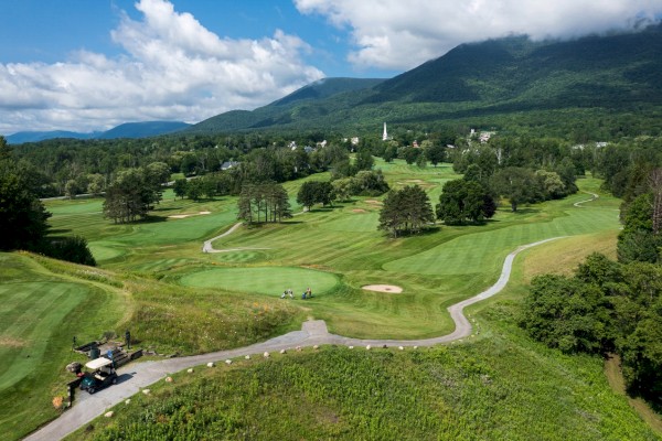 The image shows a scenic golf course with lush green fairways, people playing golf, golf carts, and mountains in the background under a partly cloudy sky.