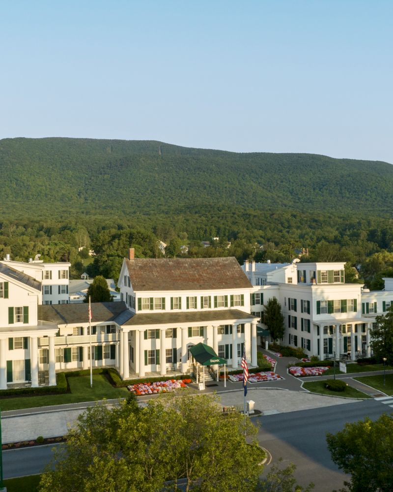 The image shows a series of white buildings, likely a hotel or resort, set against a backdrop of lush green mountains and trees, with a clear sky.