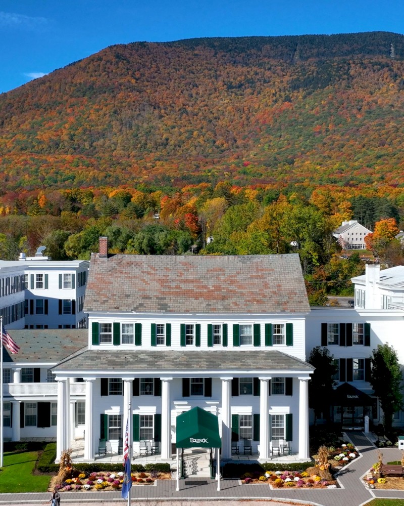 A large, white building with green shutters sits amid fall foliage, backed by a forested hillside under a clear blue sky.