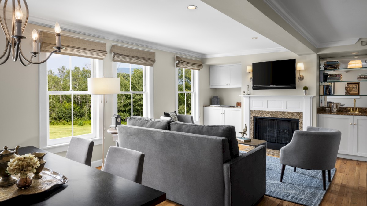 This image shows a cozy living room with a firelit fireplace, a large TV, gray sofas, a dining table, and natural light streaming in from large windows.