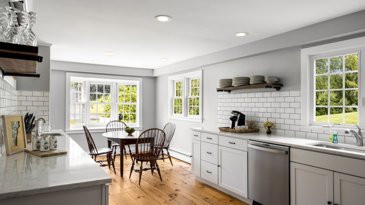 A modern kitchen with white cabinets, subway tile backsplash, wooden floors, open shelving, a dining table, and large windows providing natural light.