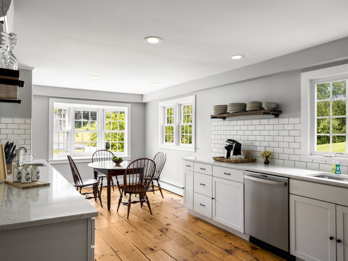A modern kitchen with white cabinets, subway tile backsplash, wooden floors, open shelving, a dining table, and large windows providing natural light.