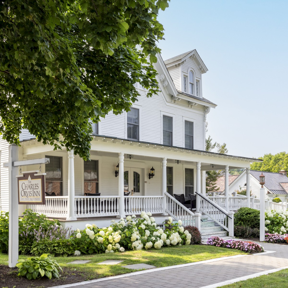 A charming white house with a wraparound porch, lush greenery, and flowers along a sidewalk, featuring a classic lamp post and a wooden sign.