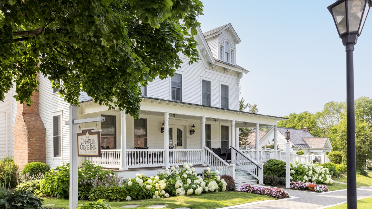 A charming white house with a wraparound porch, lush greenery, and flowers along a sidewalk, featuring a classic lamp post and a wooden sign.