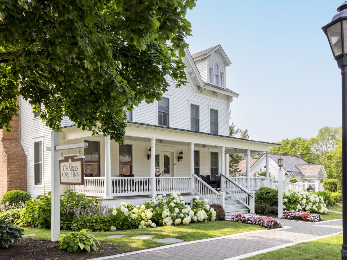 A charming white house with a wraparound porch, lush greenery, and flowers along a sidewalk, featuring a classic lamp post and a wooden sign.