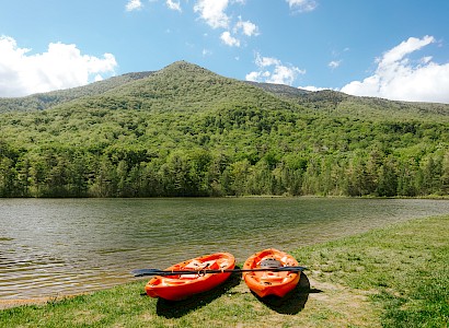 Two orange kayaks and a paddle lie on grass beside a serene lake with a forested mountain in the background under a blue sky.