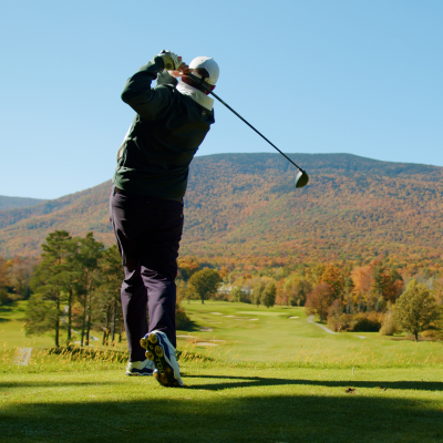 A person is playing golf on a lush course with a scenic backdrop of tree-covered hills and a clear blue sky, suggesting a beautiful autumn day.