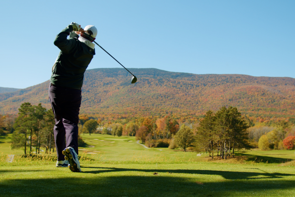 A person is playing golf on a lush course with a scenic backdrop of tree-covered hills and a clear blue sky, suggesting a beautiful autumn day.