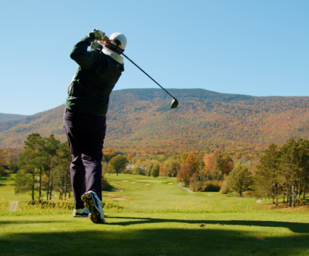 A person is playing golf on a lush course with a scenic backdrop of tree-covered hills and a clear blue sky, suggesting a beautiful autumn day.