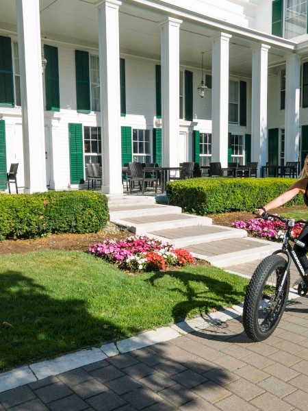 A person is riding a fat-tire bicycle on a paved path next to a large, white columned building with green shutters, surrounded by flowers and greenery.