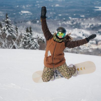 A person in winter gear and a helmet is kneeling in the snow with a snowboard, raising their arms in excitement against a snowy landscape backdrop.