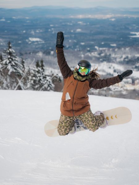 A person in winter gear and a helmet is kneeling in the snow with a snowboard, raising their arms in excitement against a snowy landscape backdrop.