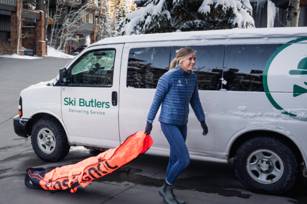 A person is walking next to a Ski Butlers delivery van, carrying a bright orange ski bag outside in a snowy area.