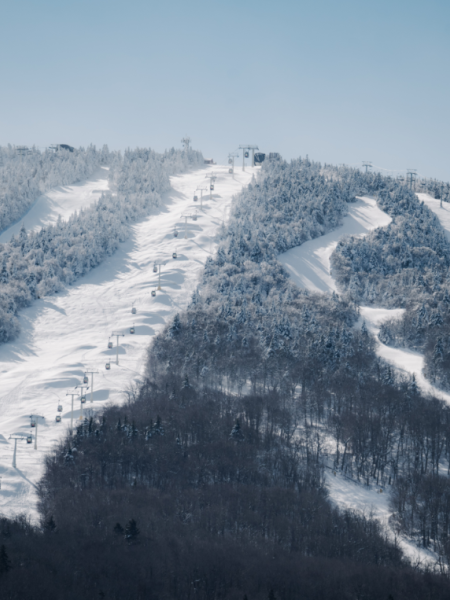 A snowy mountain ski resort with trails and ski lifts under a clear blue sky, surrounded by snow-dusted trees on both sides of the slopes.