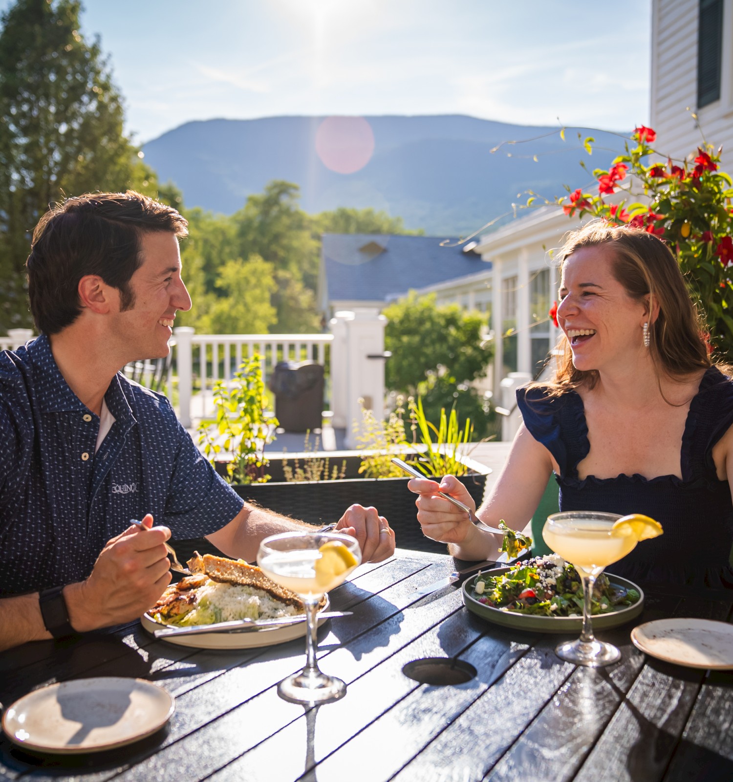 A couple enjoying a meal outdoors, surrounded by flowers, sunlight, and a scenic mountain view in the background.